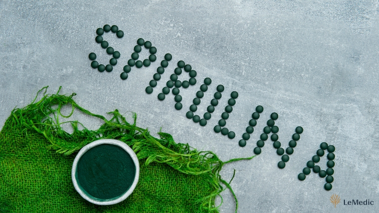 Nádpis 'Spirulina' vytvorený z tabliet spiruliny na šedom pozadí s miskou plnou prášku a zelenou tkaninou, zdôrazňujúc jej prírodný pôvod a výživové benefity.